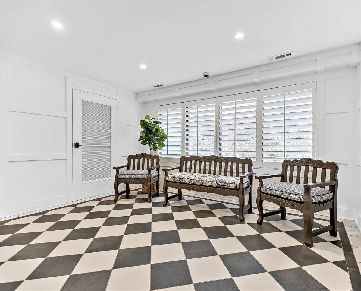 Una habitación con piso a cuadros, dos bancos de madera con asientos acolchados, una planta en maceta y paredes blancas con una ventana con persianas.