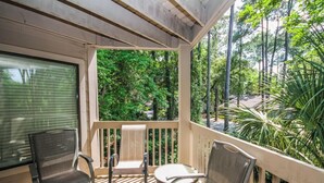A covered outdoor area with a wooden deck, two chairs, a small table, and a view of a forested area. - Lighthouse Tennis by Hilton Head Retreats (Hilton Head Island)
