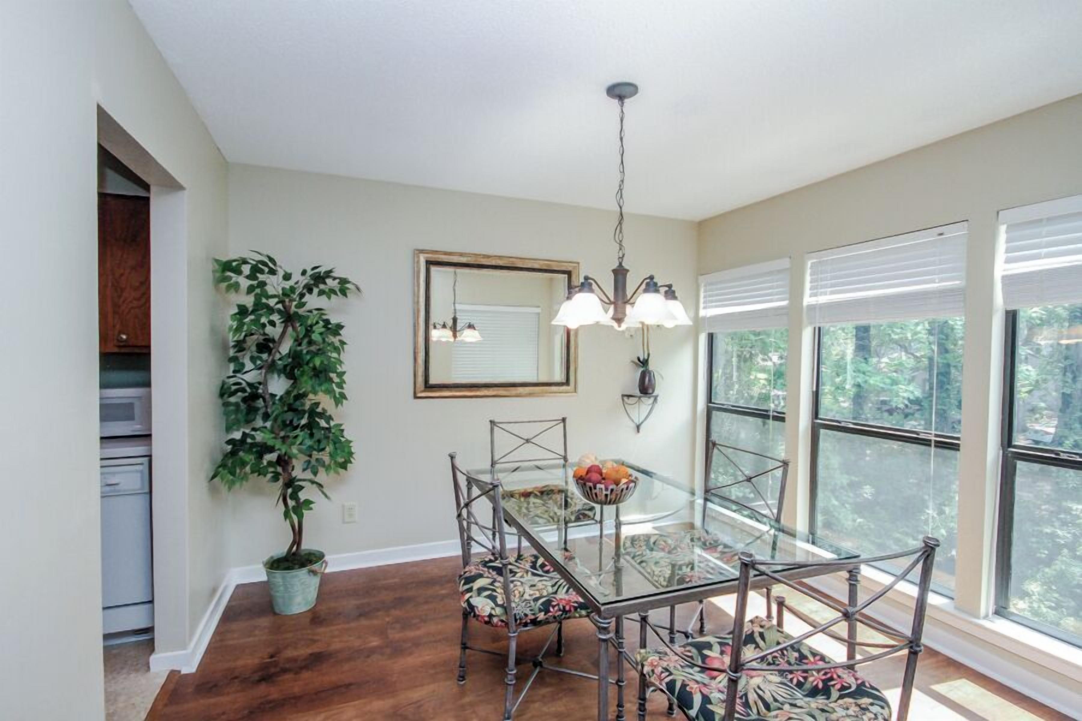 A dining area with a glass table, metal chairs, a chandelier, a potted plant, and large windows.