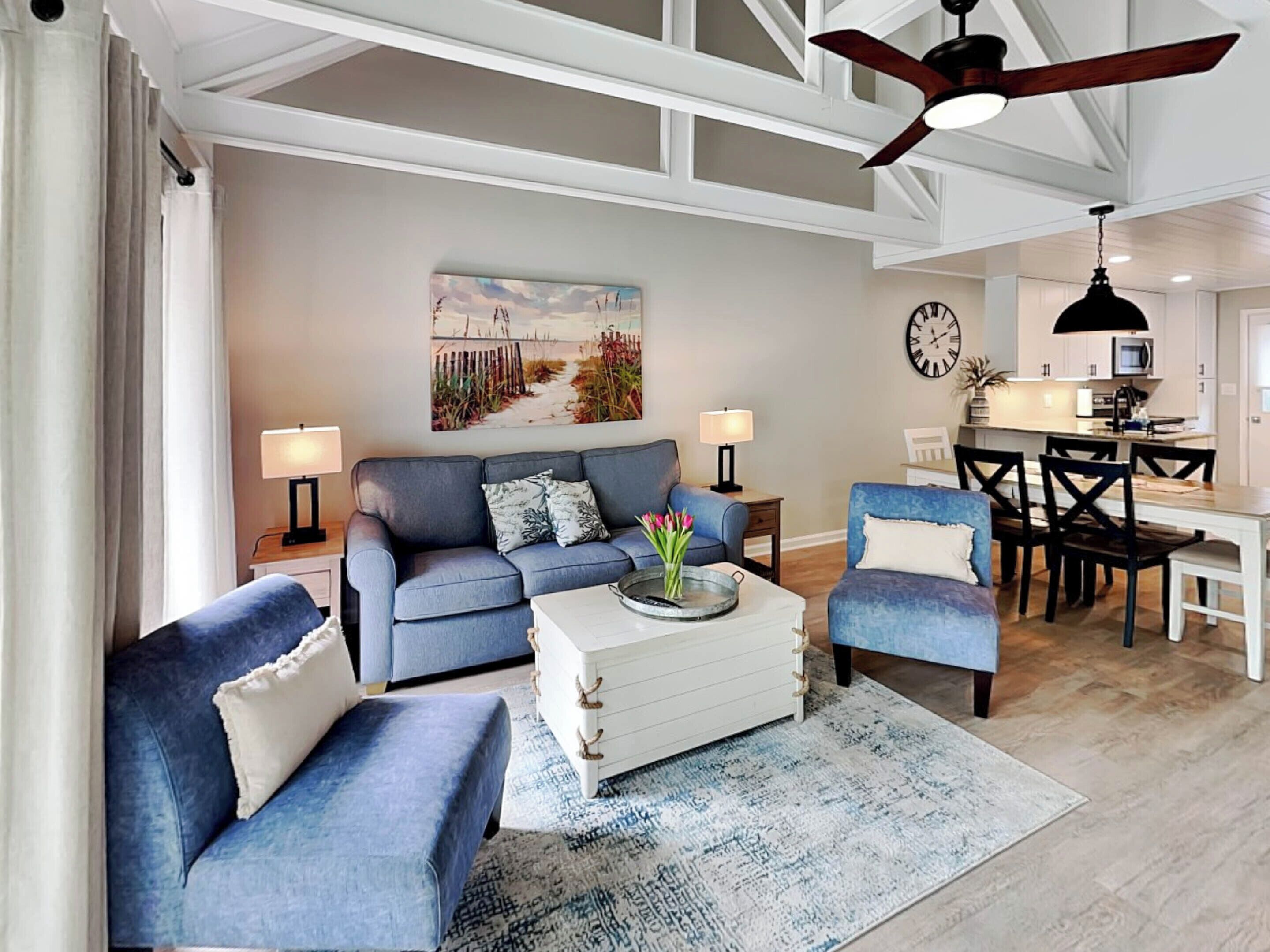 A modern living room with a blue sofa set, a coffee table, and a dining area with a kitchen in the background.