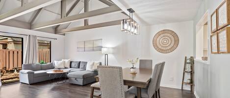 A spacious living room with a dining area, featuring a high ceiling with exposed beams, a large window, and a decorative wall piece.