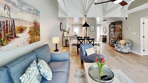 A spacious living room with a large painting, a sofa, a coffee table with flowers, and a ceiling fan. - Queens Grant by Hilton Head Retreats (Hilton Head Island)