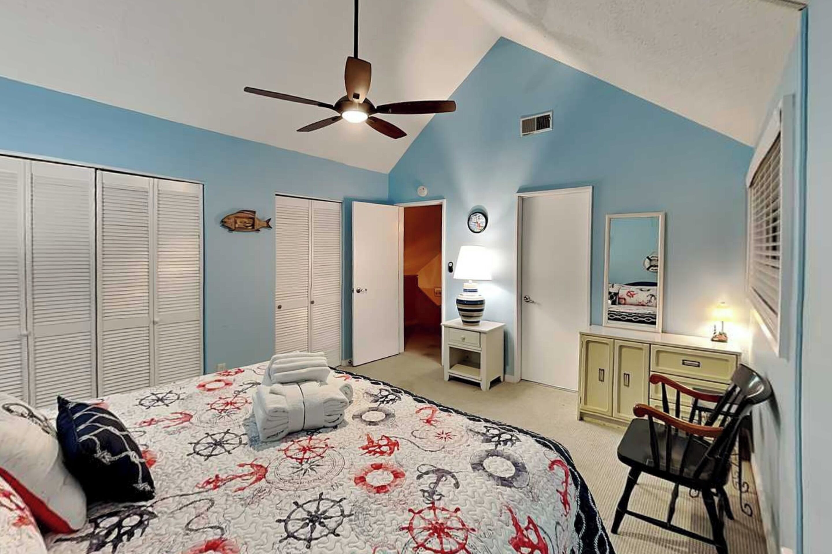 A bedroom with a bed, a ceiling fan, a dresser, a chair, and a door leading to another room.