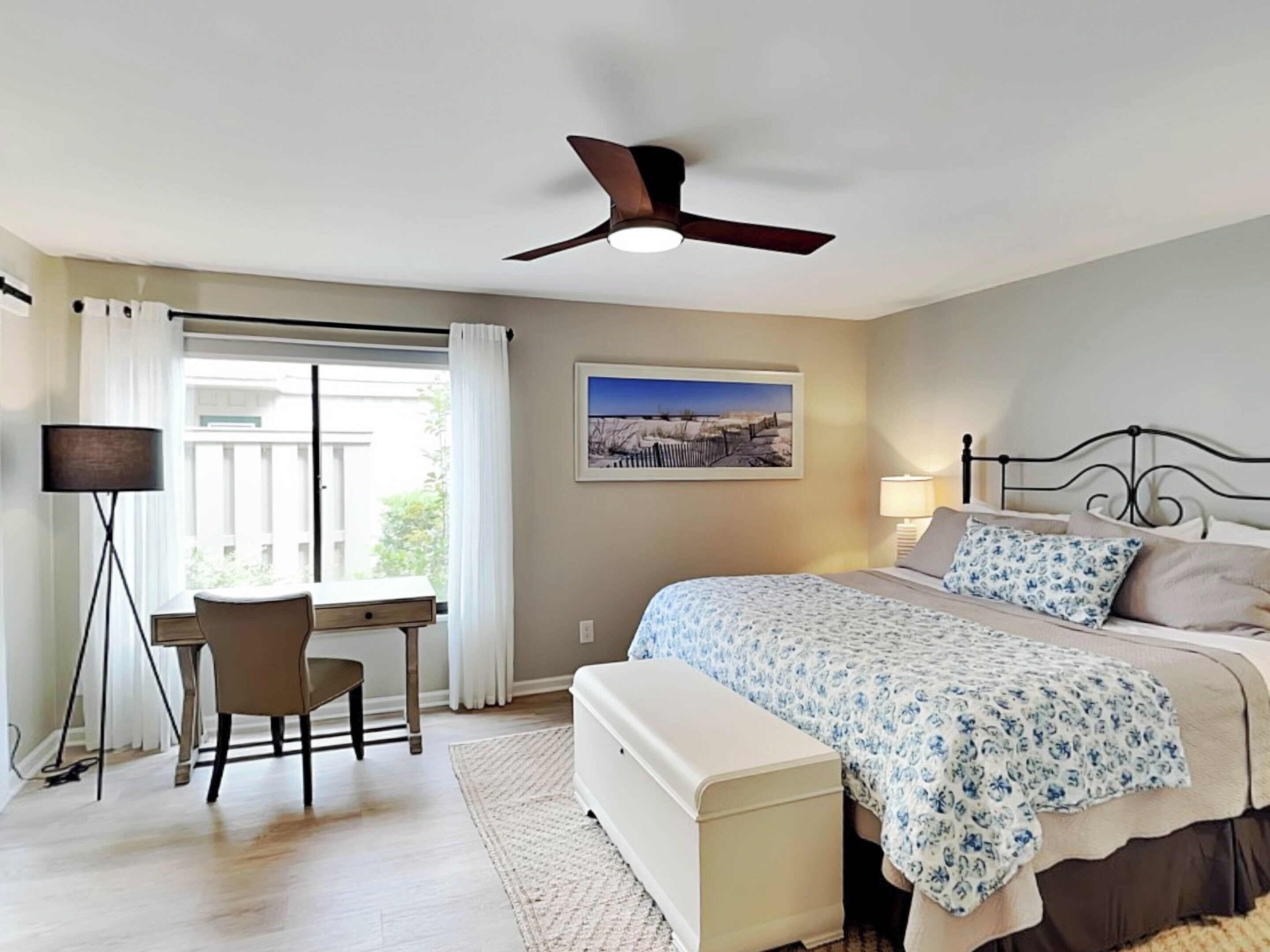 A bedroom with a bed, a desk, a chair, a ceiling fan, and a window with curtains.