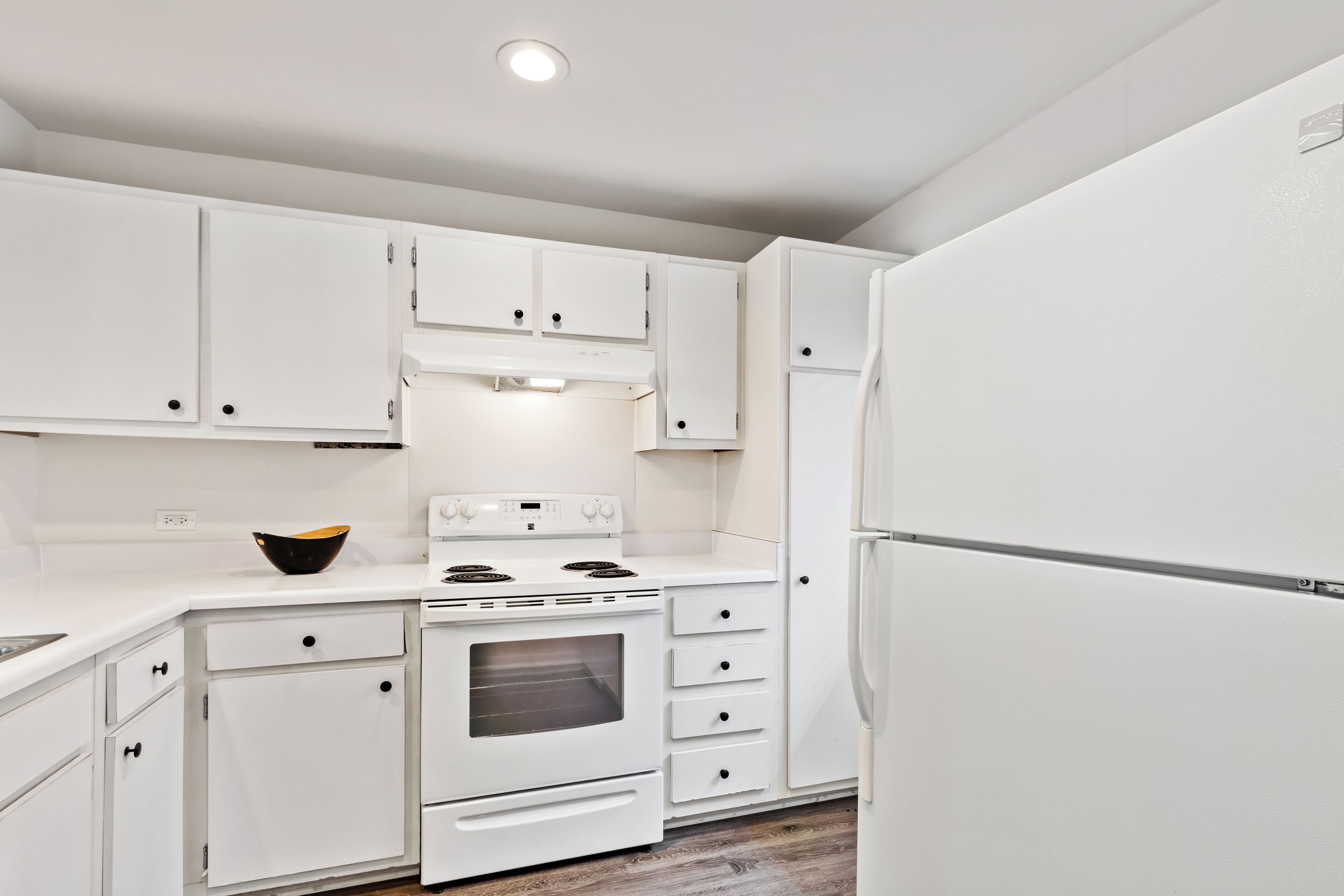 A kitchen with white cabinets, a stove, a refrigerator, and a bowl on the counter.