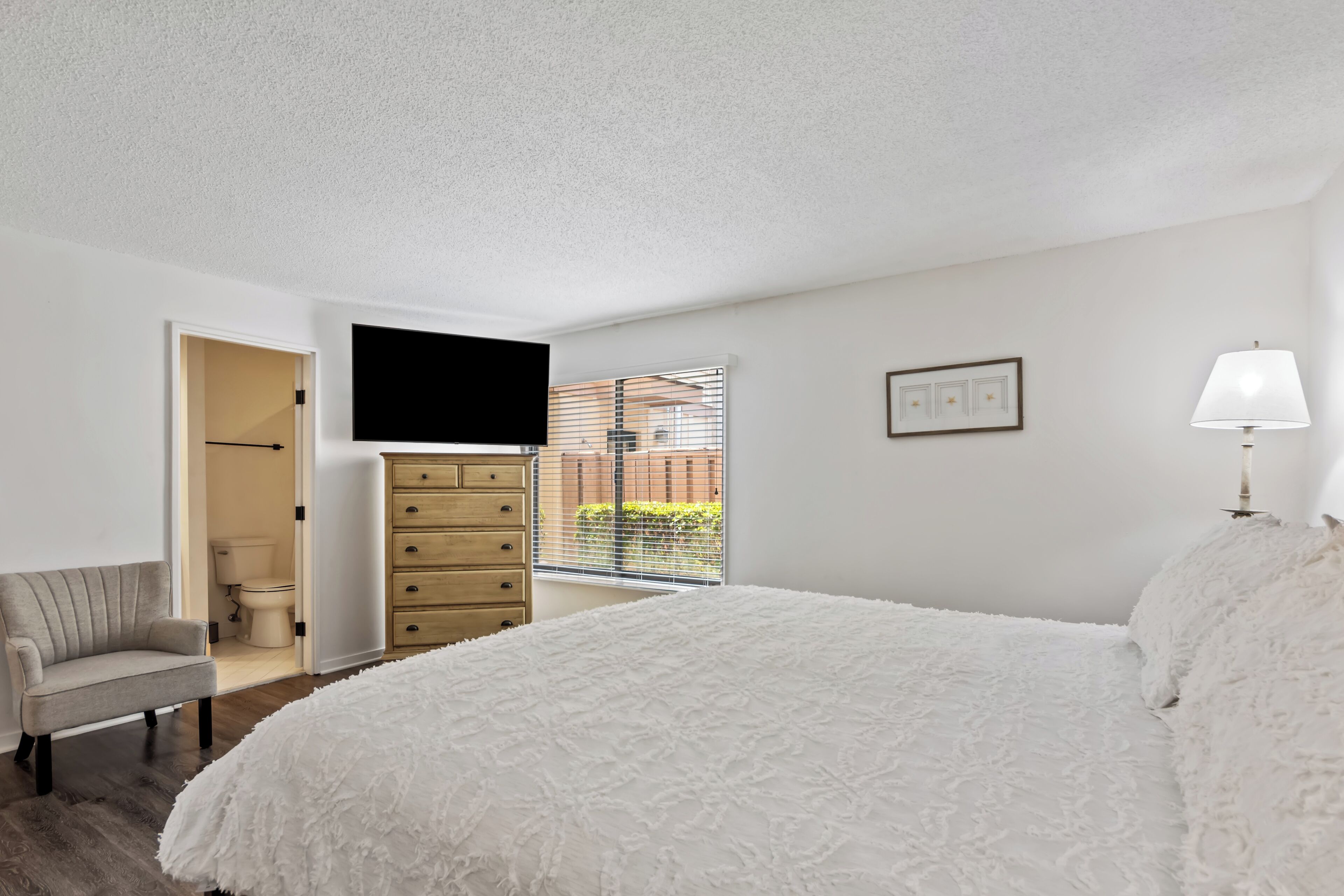 A bedroom with a large bed, a chair, a dresser, a TV mounted on the wall, and a window with blinds.