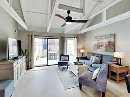 A spacious living room with a large flat-screen TV, a ceiling fan, a sofa, a coffee table, and a painting on the wall.