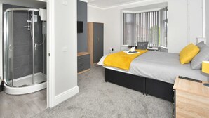 Quarto moderno com cama, mesa de cabeceira, persianas e área de chuveiro.