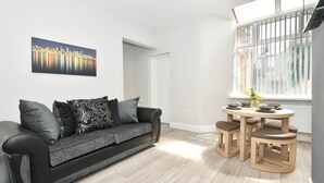 Uma sala de estar moderna com um sofá cinzento, uma pintura com vista para a cidade, uma mesa de jantar de madeira com bancos e uma janela com persianas.