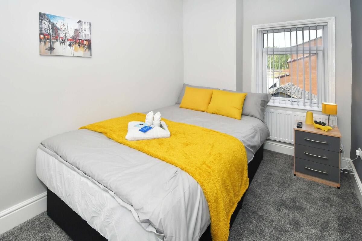 Un dormitorio ordenado con cama, mesita de noche, radiador y ventana con barrotes.