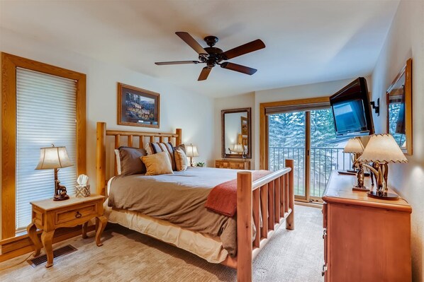 A bedroom with a bed, a ceiling fan, a television, and a balcony with a view of trees.