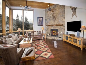 A cozy living room with a stone fireplace, leather furniture, and a large window offering a view of trees.