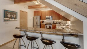 A modern kitchen with a central island, bar stools, wooden cabinets, stainless steel appliances, and a wooden ceiling beam. - Tyrolean 6 (Vail)
