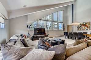 A spacious living room with a large window, a flat-screen TV, a dining area, and a modern art piece on the wall.