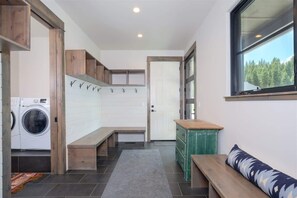 A laundry room with a washing machine, benches, a green dresser, and a window with a view of trees. - Luxury Big Sky Mountain Farmhouse (Big Sky)