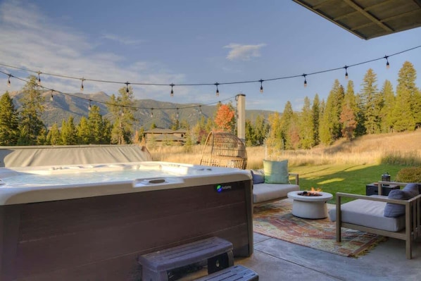 A hot tub with a view of mountains and trees, outdoor seating, and string lights. - Luxury Big Sky Mountain Farmhouse (Big Sky)