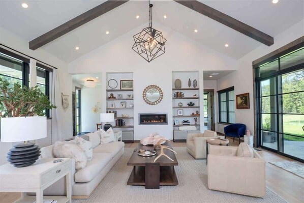 A spacious living room with a high ceiling, a fireplace, and large windows offering a view of the outdoors.