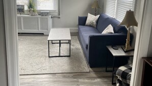 Una sala de estar con un sofá azul, una mesita de centro blanca y una lámpara. Hay una ventana con persianas y un radiador.