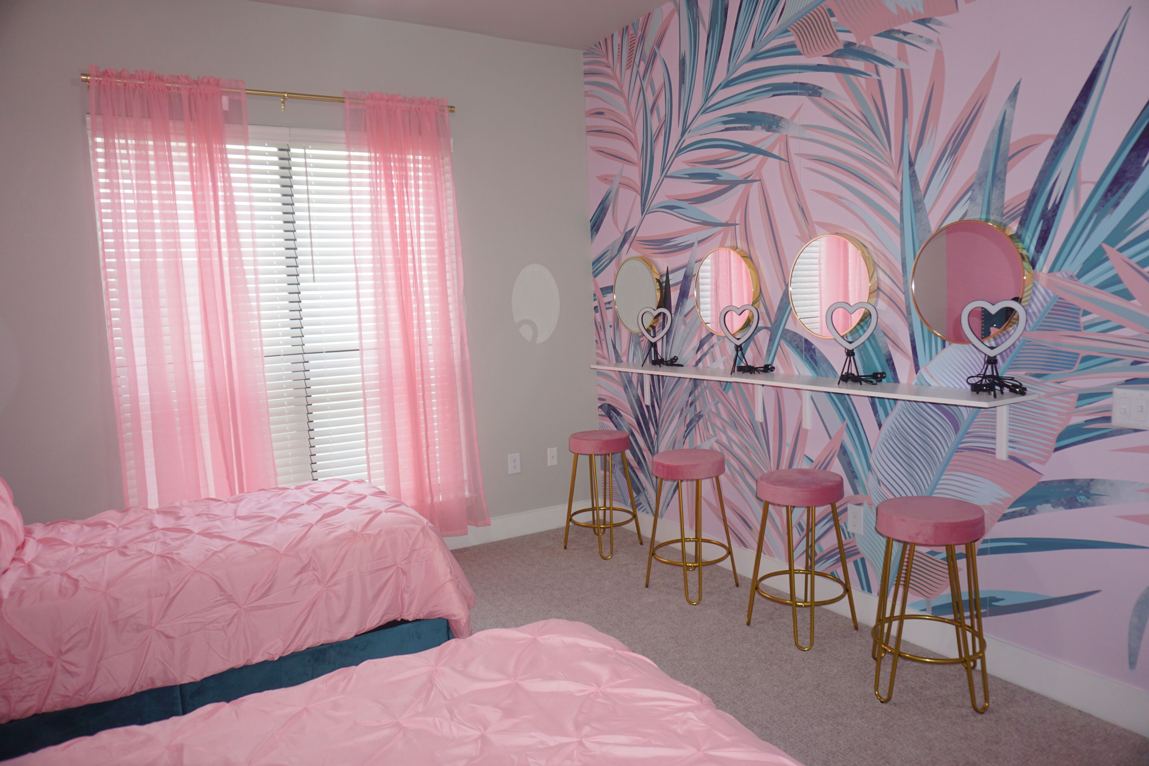 A bedroom with pink bedding, a vanity with mirrors, and a tropical leaf mural.