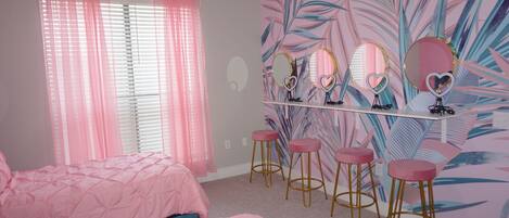 A bedroom with pink bedding, a vanity with mirrors, and a tropical leaf mural.