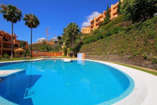 A swimming pool surrounded by palm trees and a hillside with buildings. - Apartment on the golf course (ANDALUCIA)