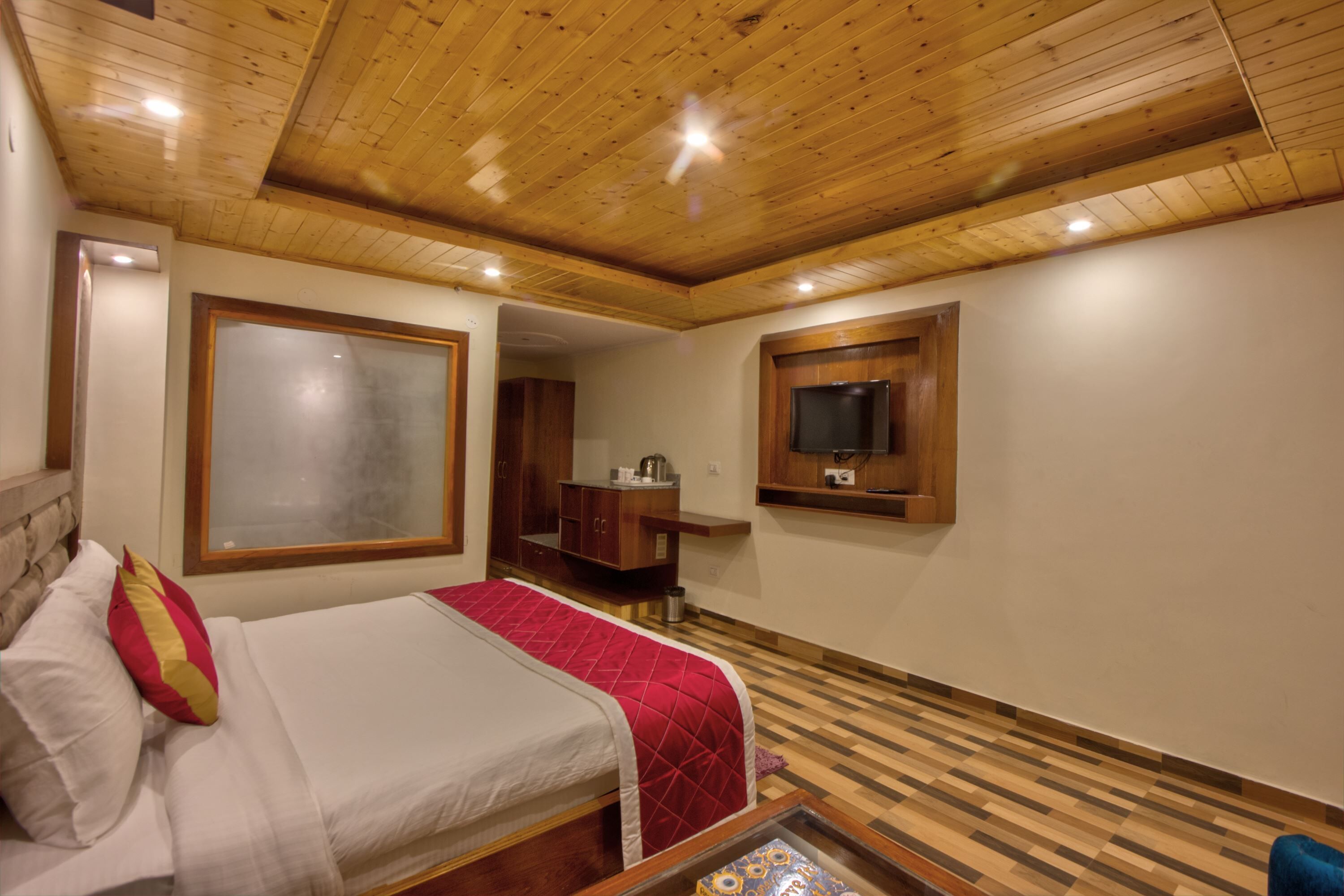 Una habitación de hotel con techo de madera, una cama con manta roja, un televisor montado en la pared y suelo de madera.
