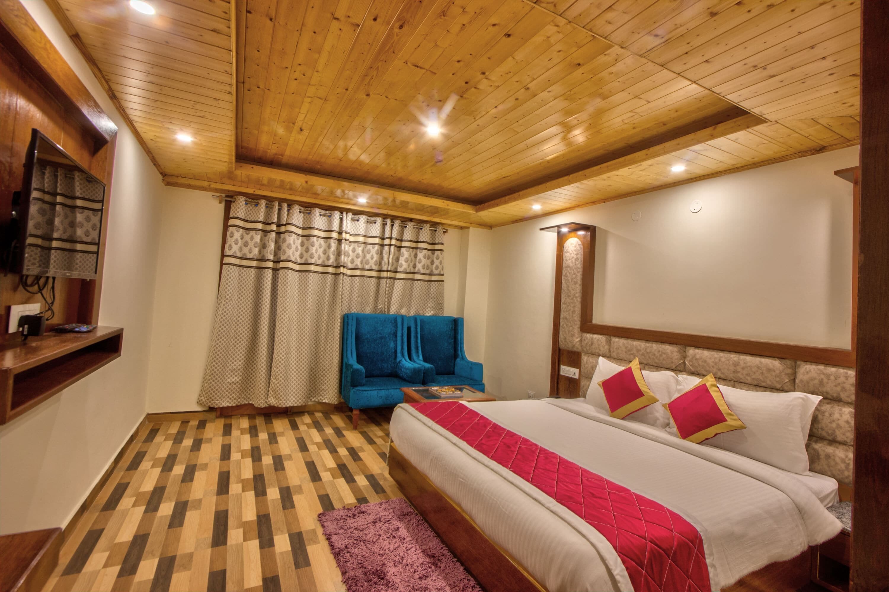 Un dormitorio con techo de madera, cama con ropa de cama roja y blanca, un sillón azul y suelo a cuadros.