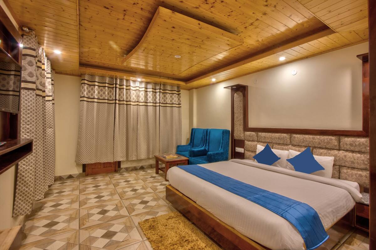 A bedroom with a wooden ceiling, a large bed with blue bedding, patterned curtains, and a blue chair.