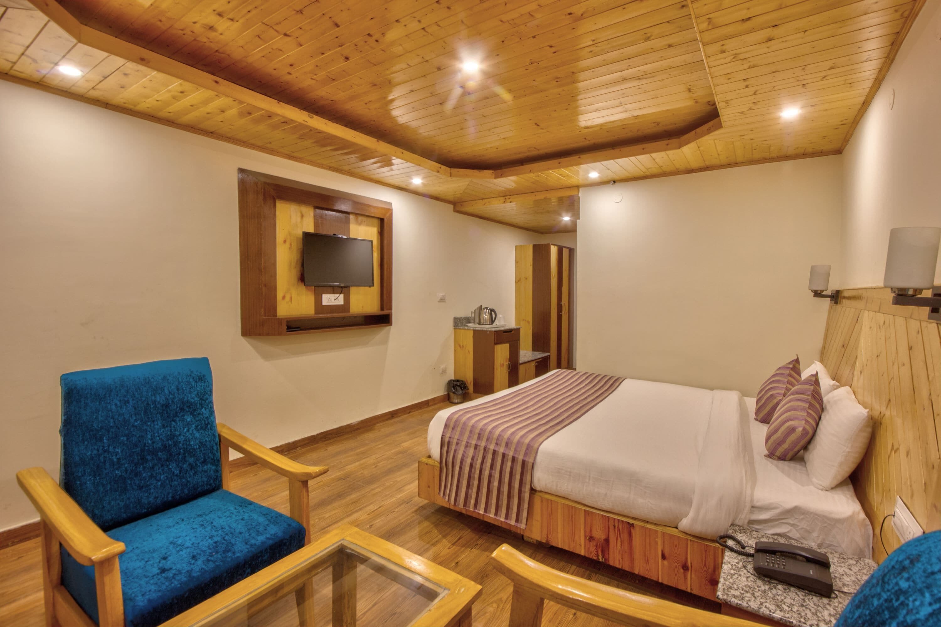 A bedroom with a wooden ceiling, a bed with a white and purple striped blanket, a blue chair, a wooden nightstand, and a television mounted on the wall.