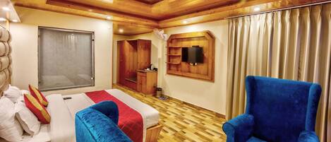 Habitación de hotel con techo de madera, cama con ropa de cama blanca y roja, sillón azul, armario de madera y televisión montada en la pared.