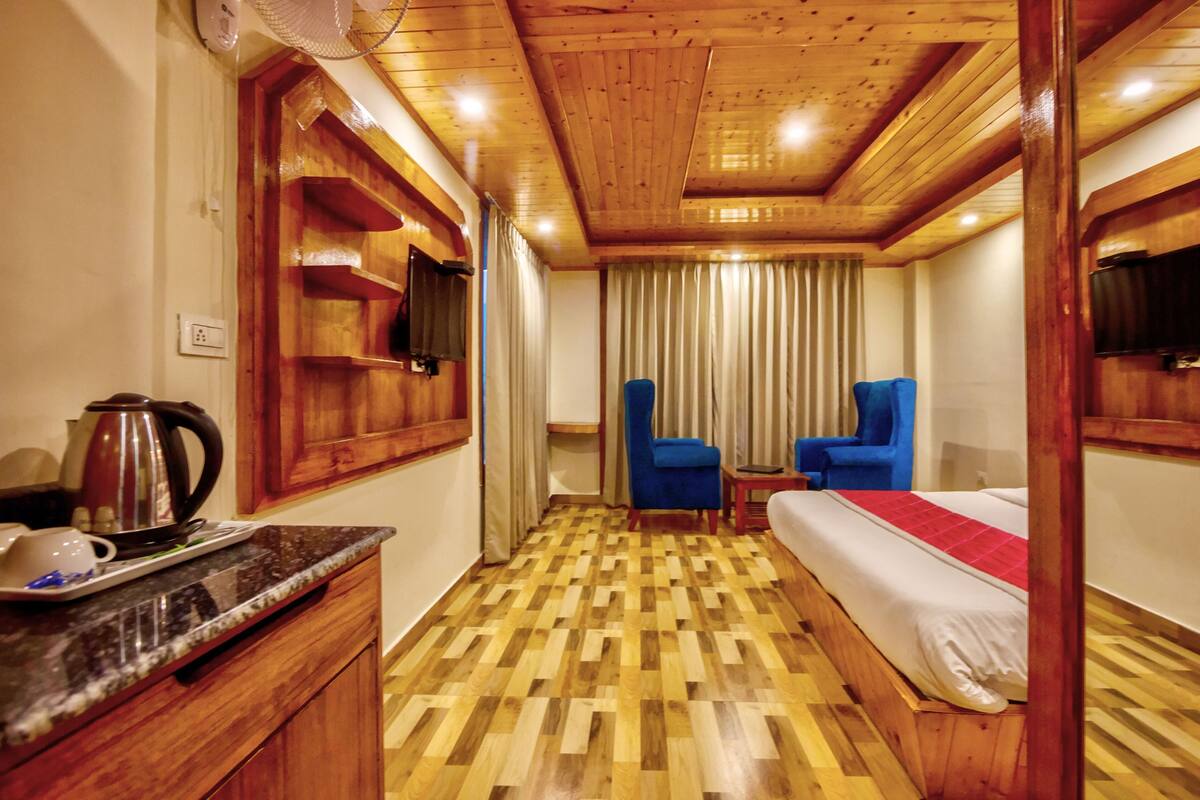 A hotel room with a wooden floor, a bed, a desk with a kettle, a chair, and a television.