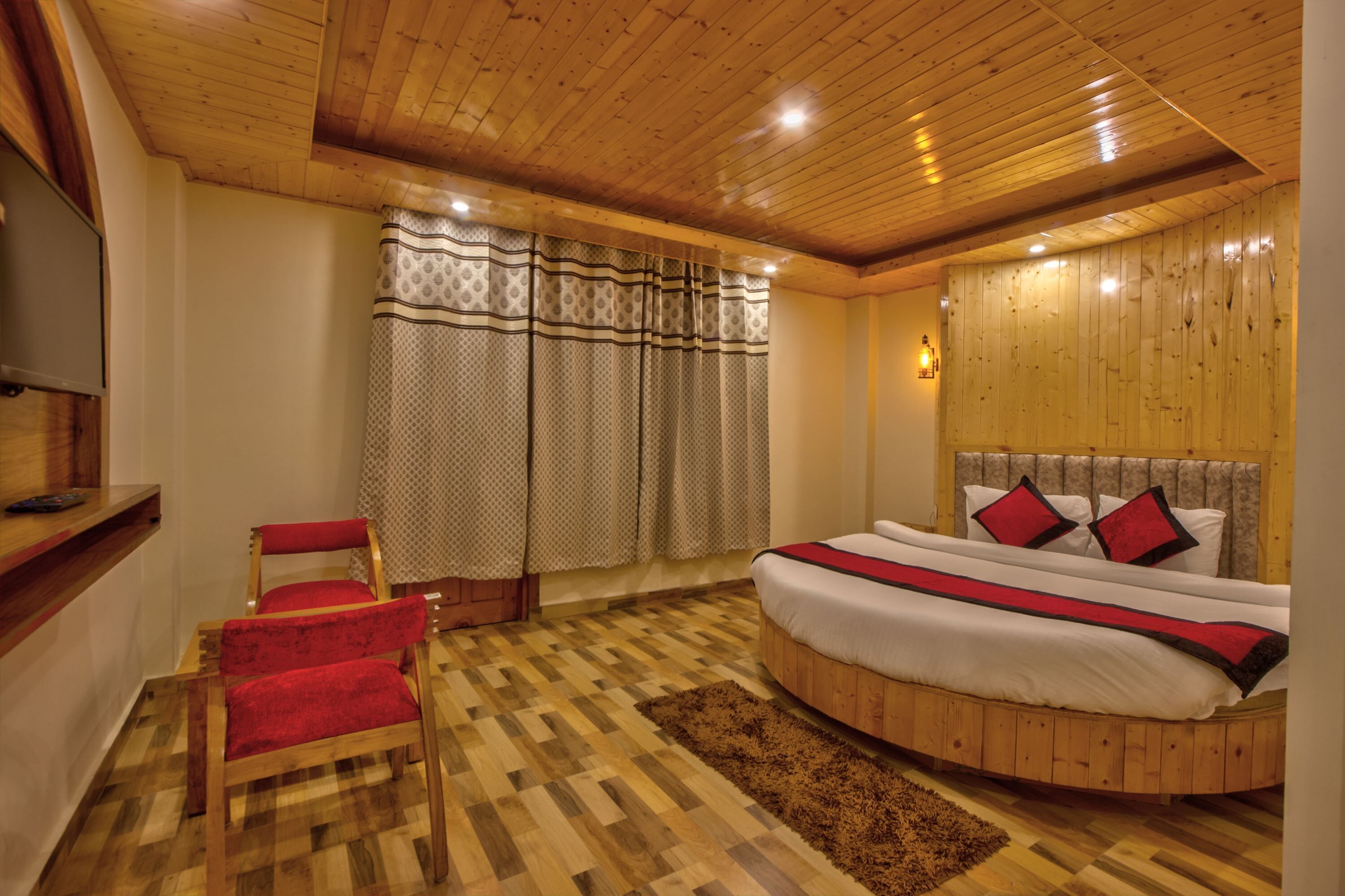 A wooden-paneled bedroom with a round bed, red chairs, and a patterned curtain.