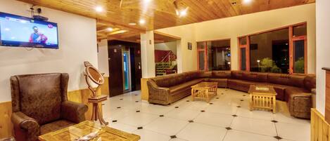 A spacious lobby with a wooden ceiling, a large TV mounted on the wall, a brown leather sofa, a glass-top coffee table, and a patterned rug.