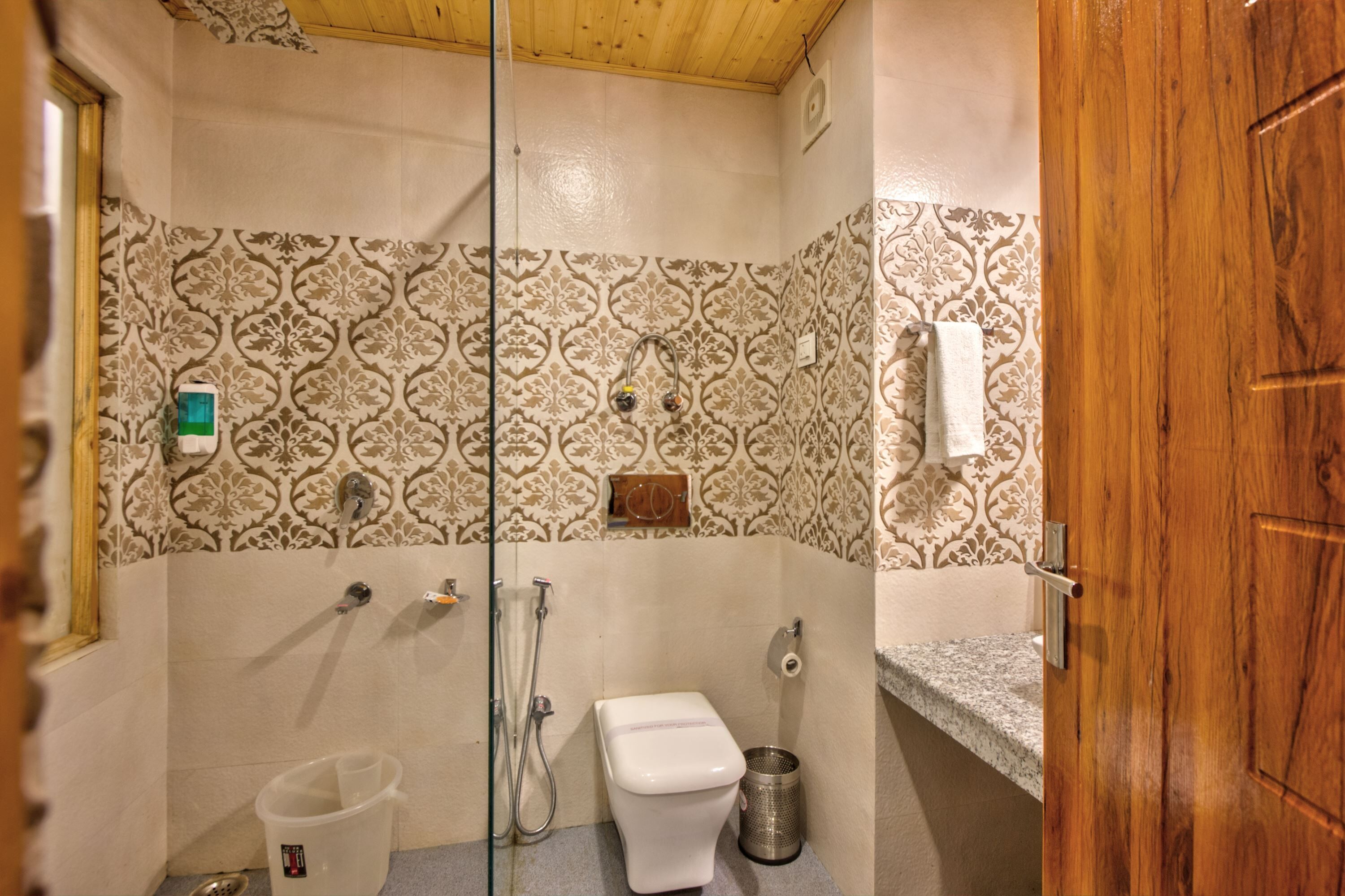 A bathroom with patterned tiles, a toilet, a shower, a trash can, and a wooden door.