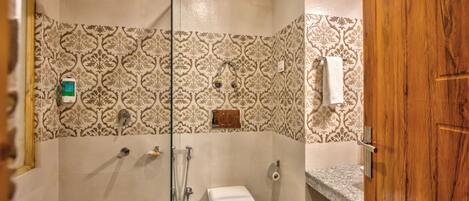 A bathroom with patterned tiles, a toilet, a shower, a trash can, and a wooden door.