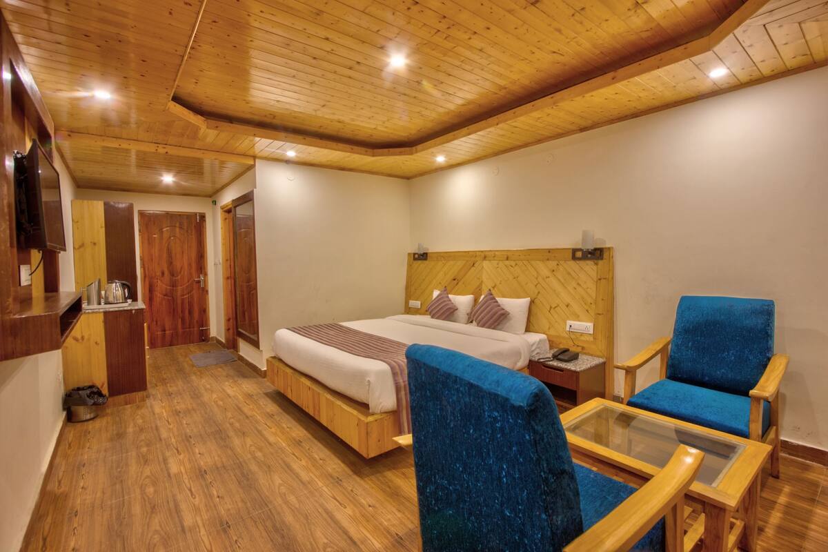 A hotel room with a wooden ceiling, a bed with a headboard, a blue armchair, a glass table, and a wooden desk with a telephone.