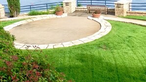 Un patio de piedra circular con un banco y plantas en macetas, con vistas a una masa de agua y una costa lejana.