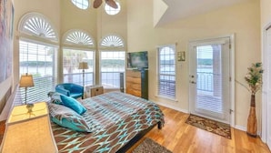 Un dormitorio con cama, ventilador de techo, televisión y vistas al exterior a través de amplios ventanales.