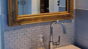 Une salle de bain avec un lavabo blanc, un miroir encadré d’or et une porte de douche.
