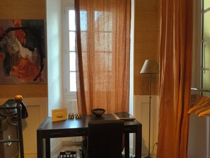 Una habitación con escritorio, silla, lámpara, un cuadro en la pared y ventana con cortinas.