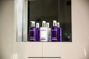 A bathroom with a mirror reflecting four bottles of Elemis products: bath & shower gel, conditioner, and shampoo.