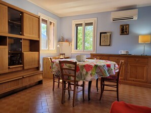 Cozinha com armários de madeira, uma mesa de jantar com toalha de mesa florida e uma janela com vista para árvores.