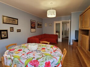 Uma sala de jantar com uma mesa redonda coberta por uma toalha florida, um sofá vermelho e um candelabro.