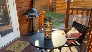 A wooden deck with a round table and two chairs, a decorative lantern, and a wooden cabin in the background. - The Cozy Cabin near Geneva on the Lake and Wineries! (Ashtabula)