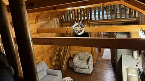 A wooden cabin interior with a loft bedroom, a staircase, a sofa, a patterned armchair, and a kitchen area. - The Cozy Cabin near Geneva on the Lake and Wineries! (Ashtabula)
