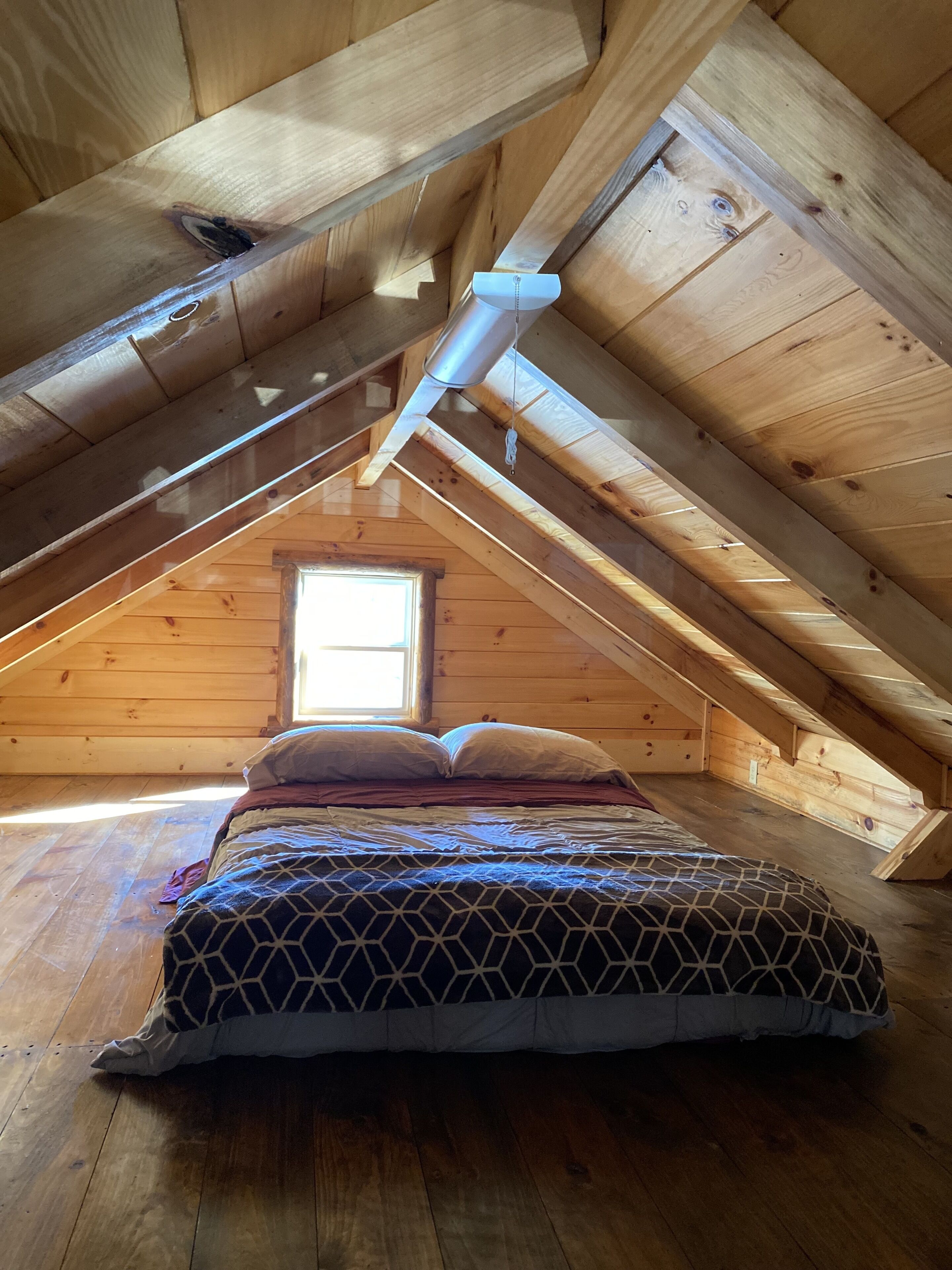 A small, cozy attic bedroom with a sloped ceiling, wooden beams, a single window, and a bed with patterned bedding.