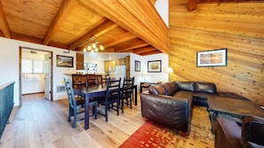 Un intérieur chaleureux avec un plafond en bois, un coin repas avec une table et des chaises, un canapé en cuir et une cuisine en arrière-plan.