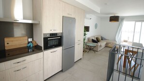 A modern kitchen with light wood cabinets, a stainless steel refrigerator, and a built-in oven. There is a dining area with a table and chairs, and a sofa in the background. - 250 - Casa Birgit (Tarifa)