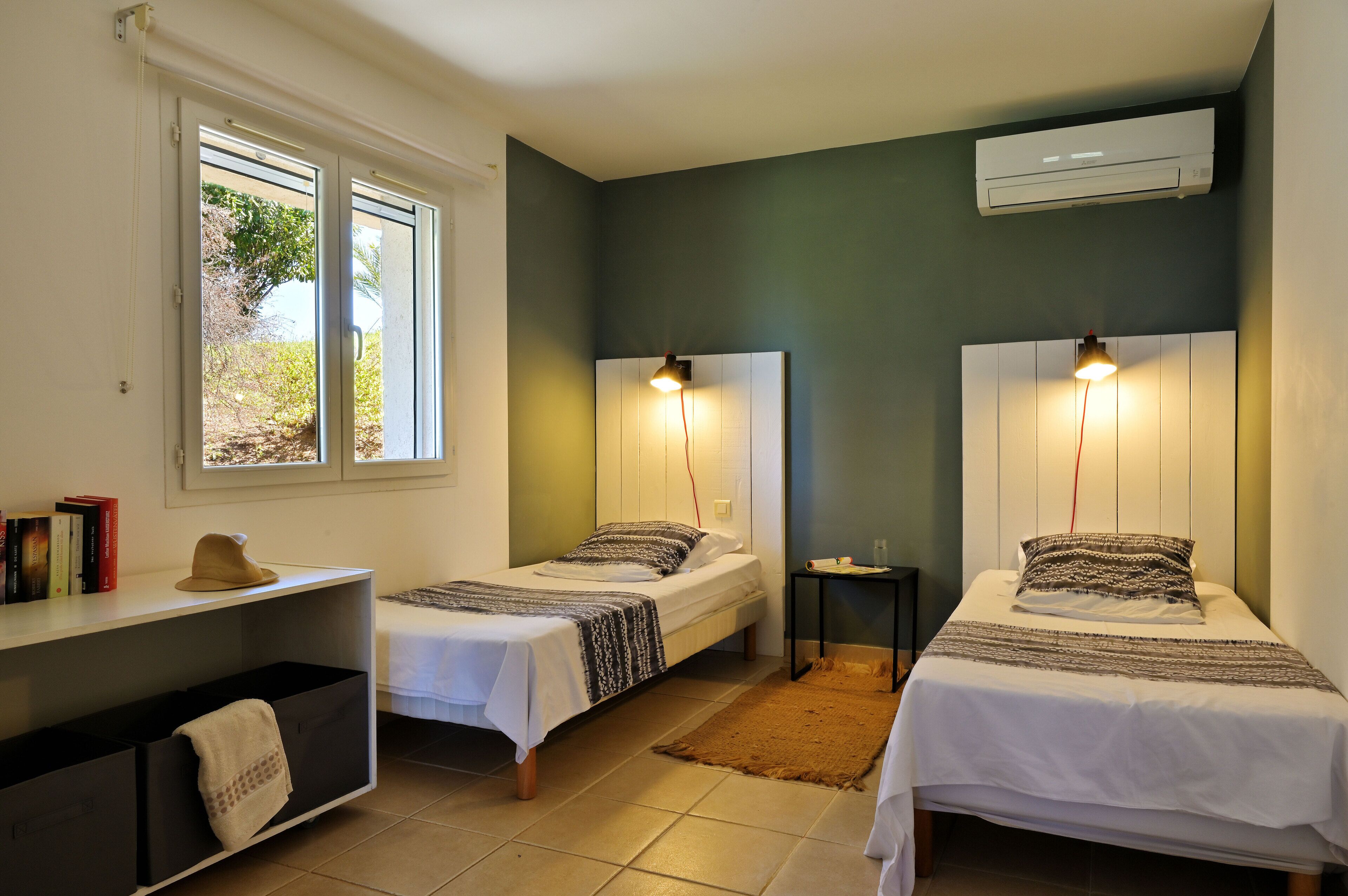 A room with two single beds, a small table, a shelf with books, and a window with blinds.