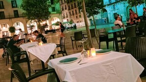 Een terras met tafels voor twee personen, omringd door bomen en een historisch pand op de achtergrond.